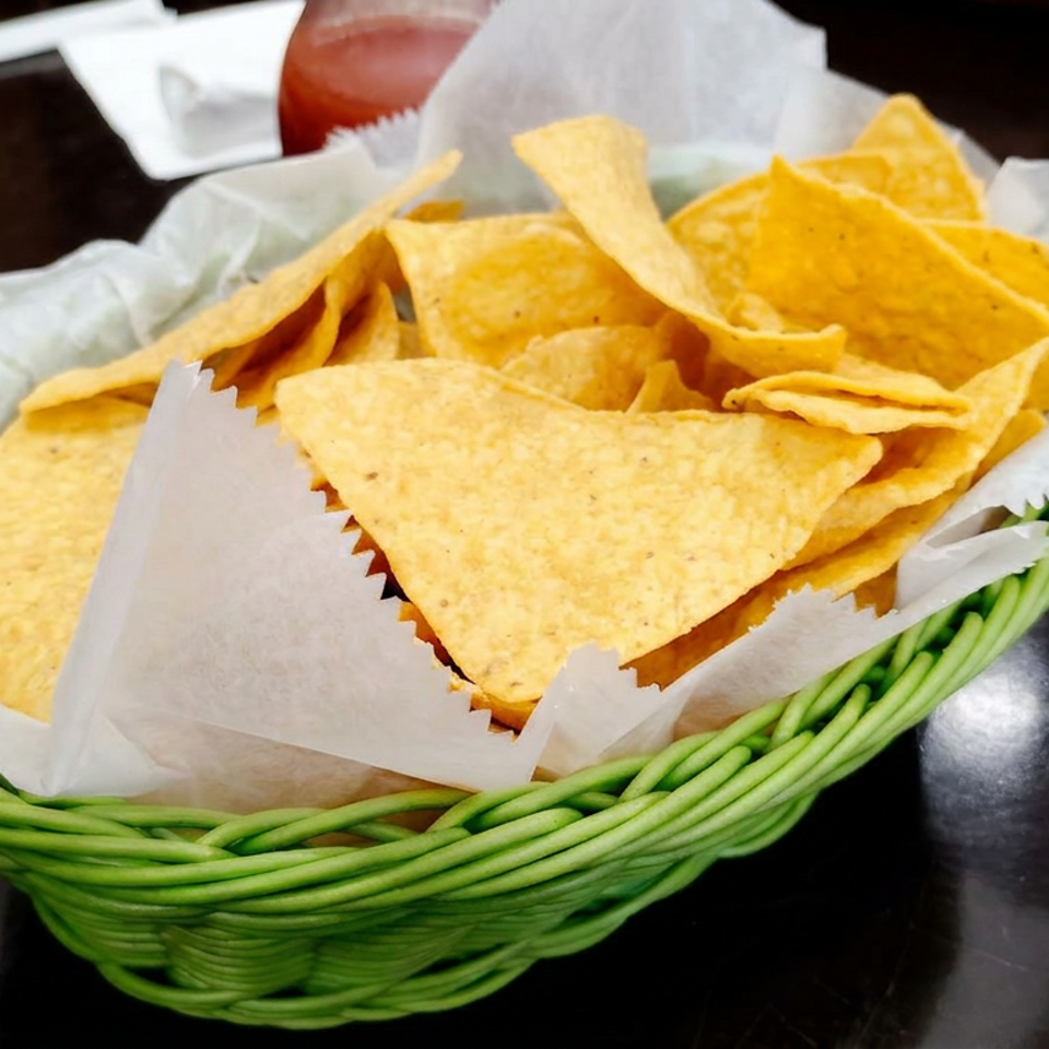 Basket of golden tortilla chips lined with paper on a restaurant table, with salsa and a diner’s hand nearby.